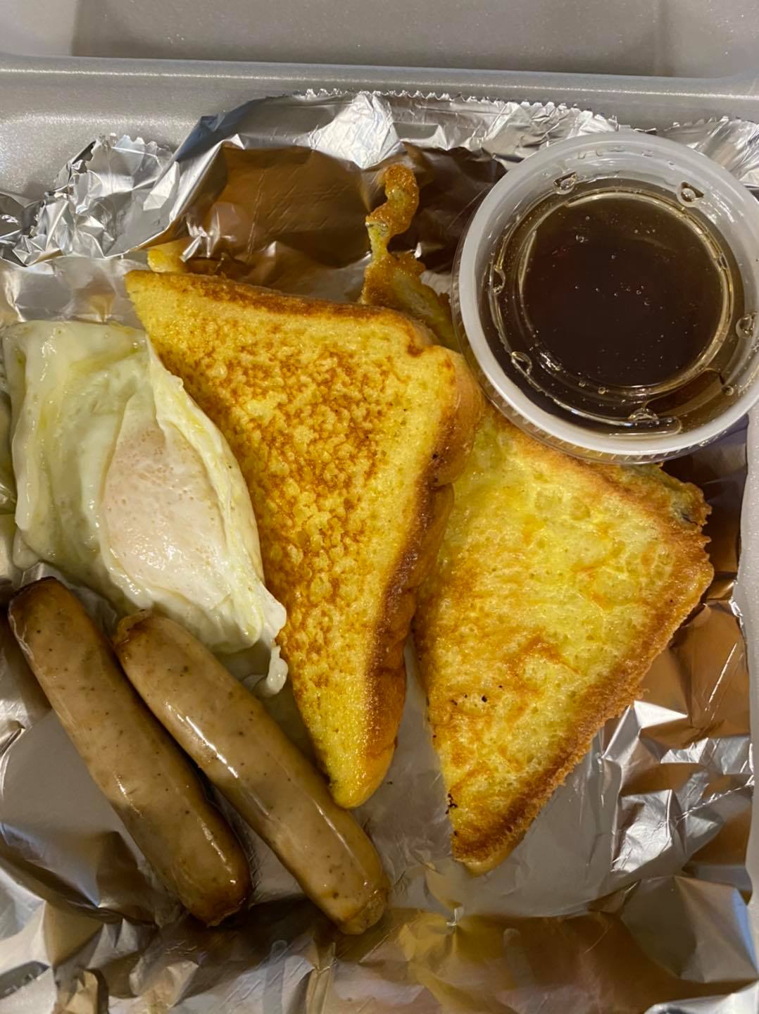 a togo container with two sausage links, egg over easy, French toast and maple syrup in a container