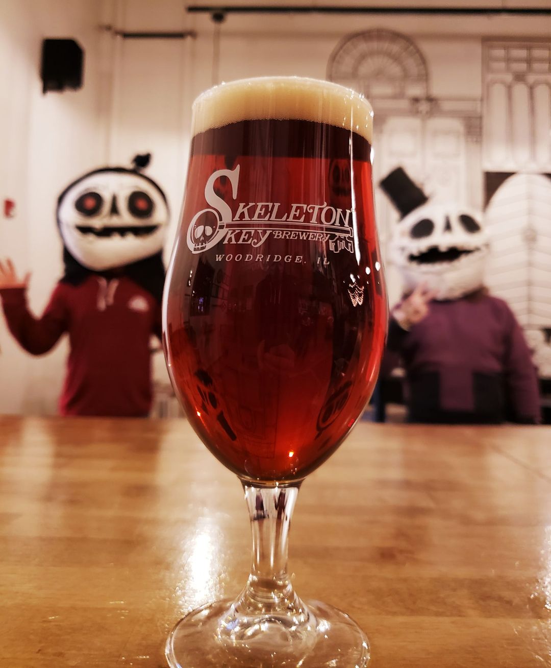 Glass of Don't Blink in the foreground from Skeleton Key Brewery, with two guys in skull head costumes in the blurry background